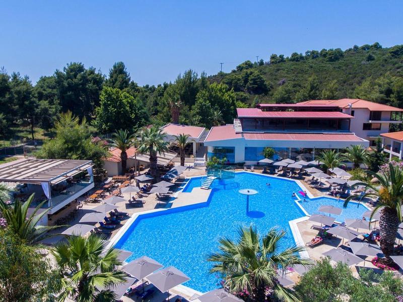 Großer blauer Pool im Resort mit Sonnenschirmen, Palmen und umliegenden Gebäuden in grüner Landschaft.