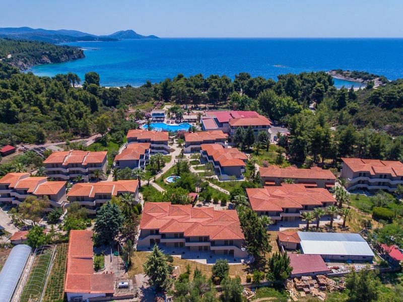 Landschaft mit zahlreichen Gebäuden und roten Dächern nahe der Küste und blauem Meer im Hintergrund.