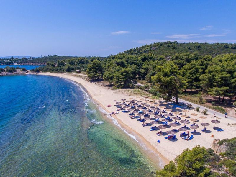 Strand mit blauem Wasser, Sonnenschirmen, Sand und bewaldeten Hügeln im Hintergrund.