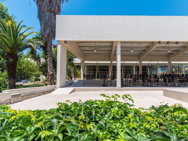 Moderner weißer Pavillon mit Stühlen, umgeben von grünen Pflanzen und Palmen bei klarem Himmel.