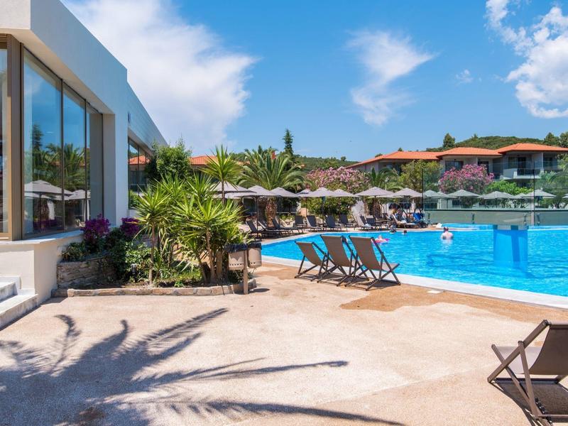 Freizeitpool mit Liegestühlen, Sonnenschirmen und mediterraner Landschaft unter blauem Himmel.