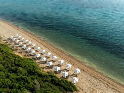 Strand mit Sand, Reihen weißer Sonnenschirme und grünem Buschwerk am Ufer eines ruhigen Meeres.