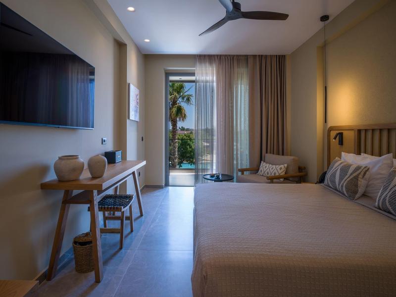 Modern hotel room with double bed, TV, fan, and view of terrace with plants.