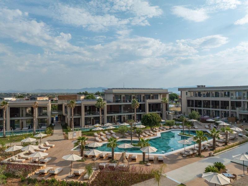 Modern hotel complex with large pool, sun loungers, and palm trees under cloudy sky.