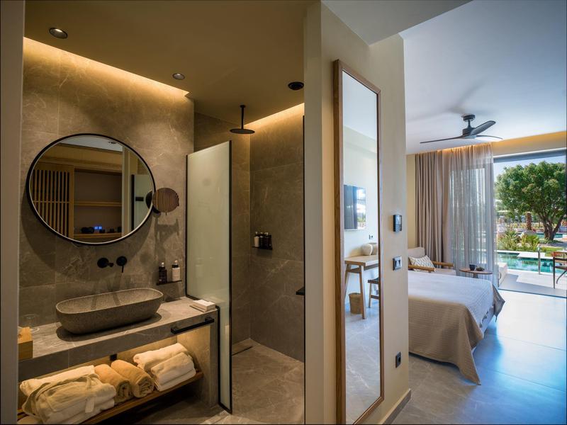Modern hotel room with bathroom and view of pool and garden through large windows.