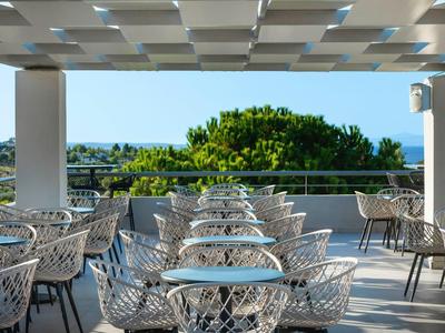 Terrassenbereich eines Hotels mit modernen Stühlen und Tischen, Blick auf grüne Bäume und blaues Meer.