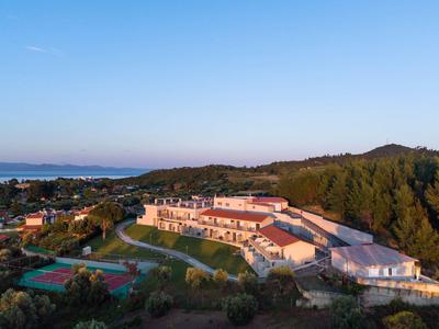 Hotel complex on a forested hill with tennis courts and sea view at sunset.