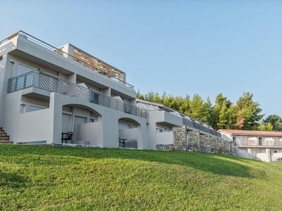 Moderní bílý hotelový komplex s balkony a zeleným trávníkem v popředí pod jasnou oblohou.