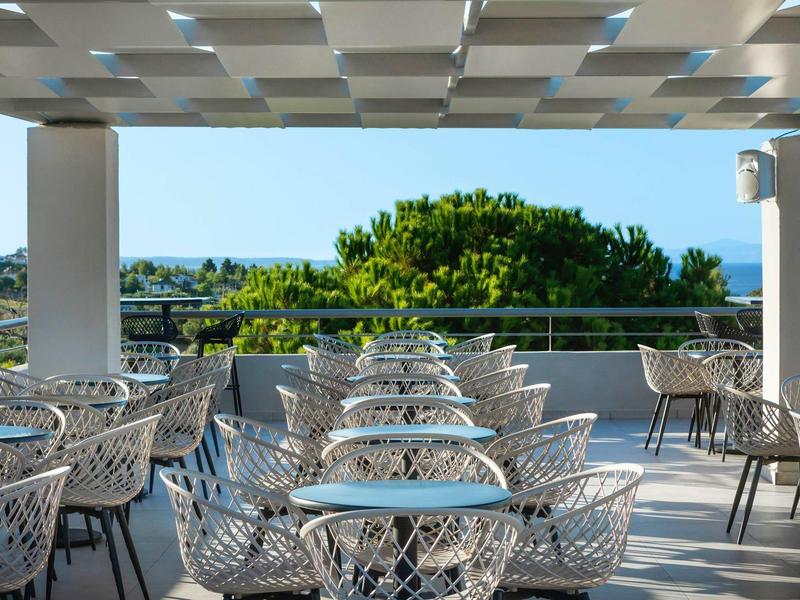 Prázdná terasa s bílými stoly a židlemi, výhled na zelené stromy a modrou oblohu.