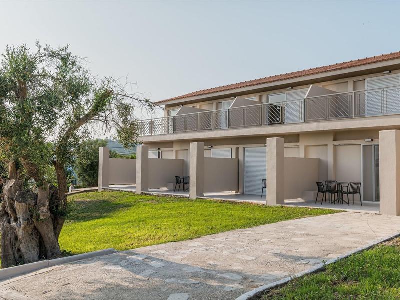 Moderní jednopatrová budova hotelu s terasou, přilehlým trávníkem a stromem v popředí.