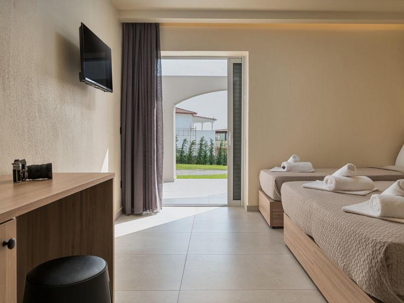 Světlejší hotelový pokoj se dvěma postelemi, TV, závěsem a otevřenými dveřmi do zahrady.