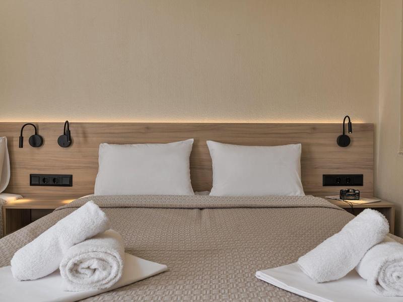 Moderní hotelový pokoj s manželskou postelí, bílými polštáři a složenými ručníky na šedém přehozu.