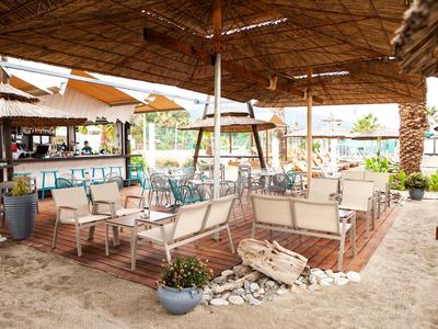 Überdachte Strandbar mit Sitzgelegenheiten und sandigem Boden, umgeben von Pflanzen.