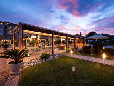 Abendliche Außenansicht eines modernen Hotels mit beleuchteter Terrasse und blau-lila Himmel.