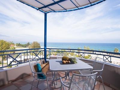 Balkon mit weißem Tisch und Stühlen, blauer Balkonbrüstung, Blick auf Meer und Himmel bei Tageslicht.