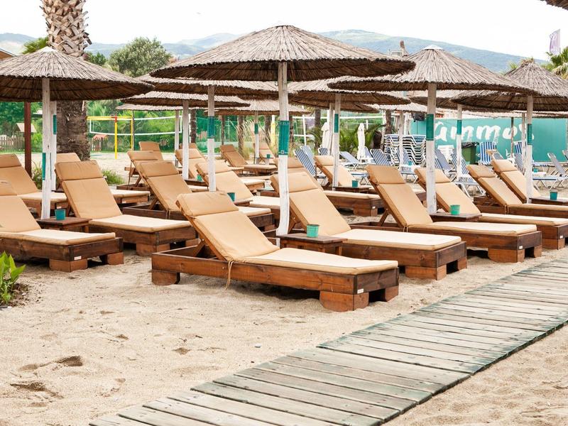 Mehrere Strandliegen mit Sonnenschirmen auf Sand neben einem Holzsteg in einer sonnigen Umgebung.