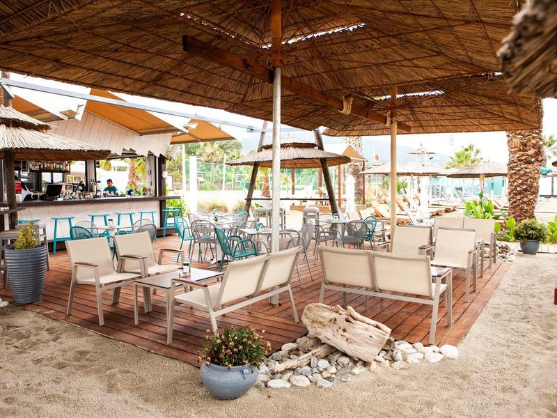 Offener Strandpavillon mit Strohdach, weißen Sitzbänken, Holzstühlen und Sandboden.