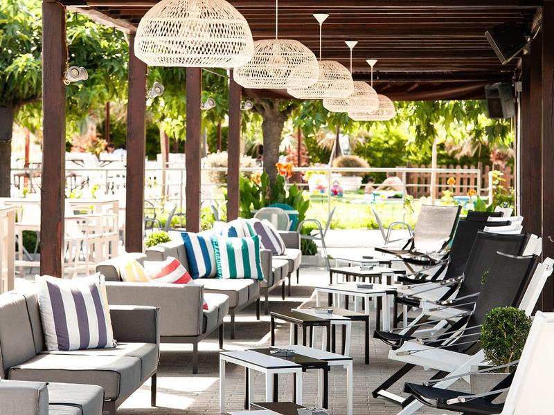 Gemütliche Außenlounge mit Sofas, Kissen und Hängelampen unter Holzdach an sonnigem Tag.