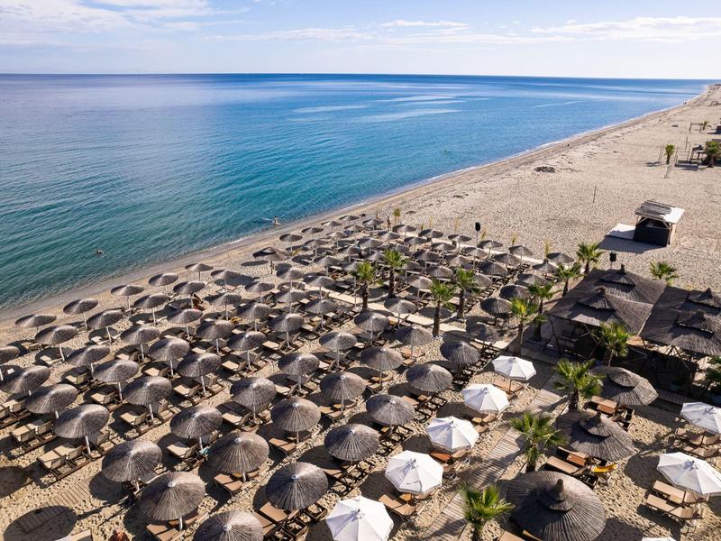 Strand mit vielen Sonnenschirmen, Sand, blauem Meer und klarem Himmel im Hintergrund.