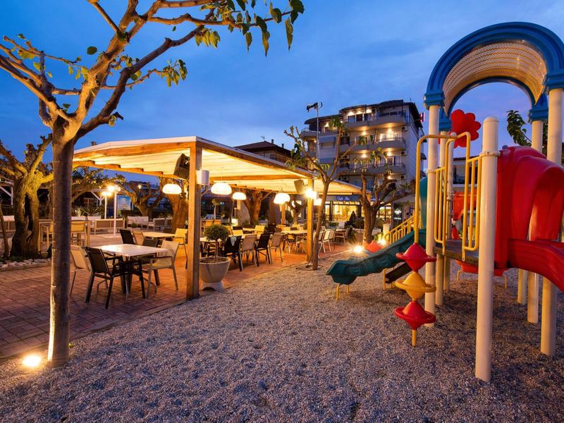 Spielplatz mit bunten Rutschen und beleuchtetem Außenrestaurant bei Dämmerung, Bäume und Gebäude im Hintergrund.