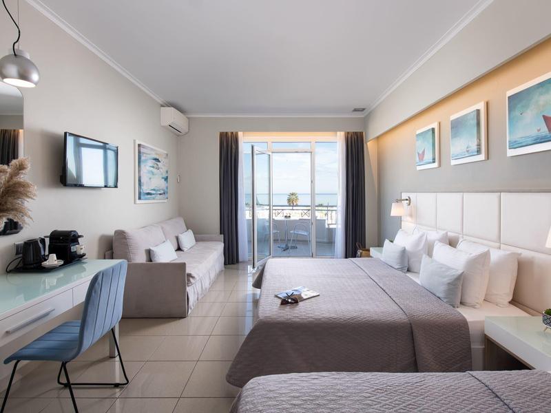 Hell eingerichtetes Hotelzimmer mit zwei Betten, Stuhl, Tisch, Sofa und Balkon mit Meerblick.