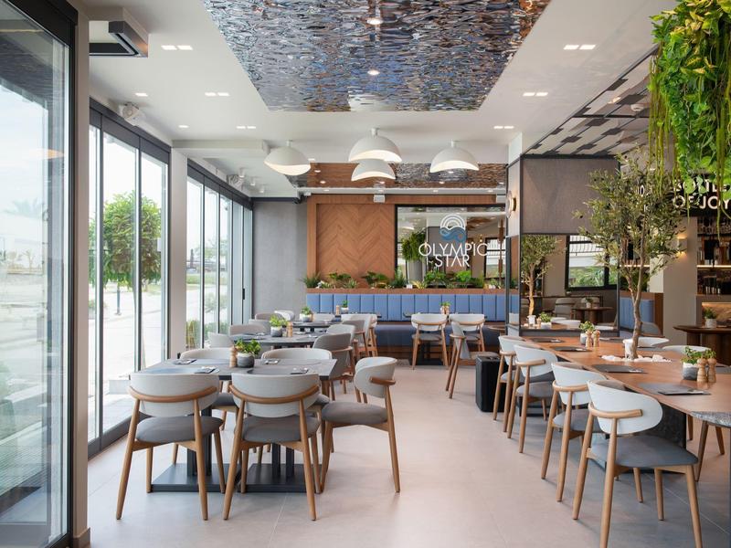 Modernes Café mit großen Fenstern, hellen Holzmöbeln, Pflanzen und Säulen mit Betonoptik.