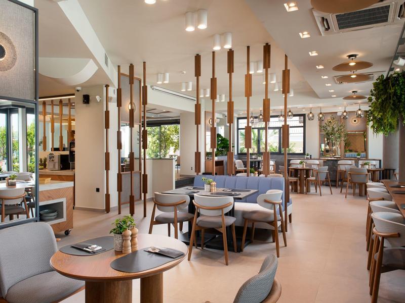 Modernes Restaurant mit heller Einrichtung, Holztischen, grauen Stühlen und großen Fenstern.