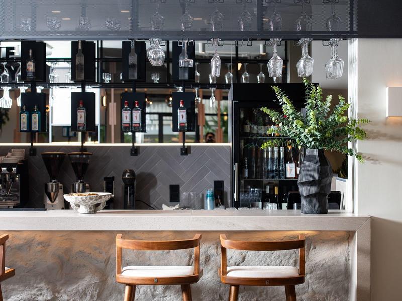 Moderner Barbereich mit Holzstühlen, Spiegelwand, Pflanzen und Regalen voller Flaschen und Gläser.