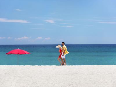 Deux personnes marchent le long d'une plage de sable blanc à côté d'un parasol rouge sous un ciel clair.