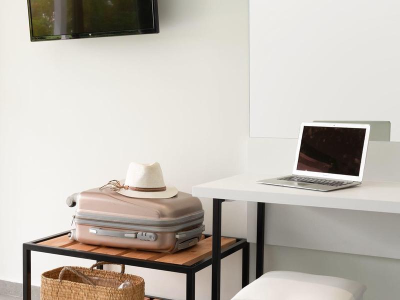A modern room with wall TV, laptop on desk, and luggage rack with suitcase and hat.
