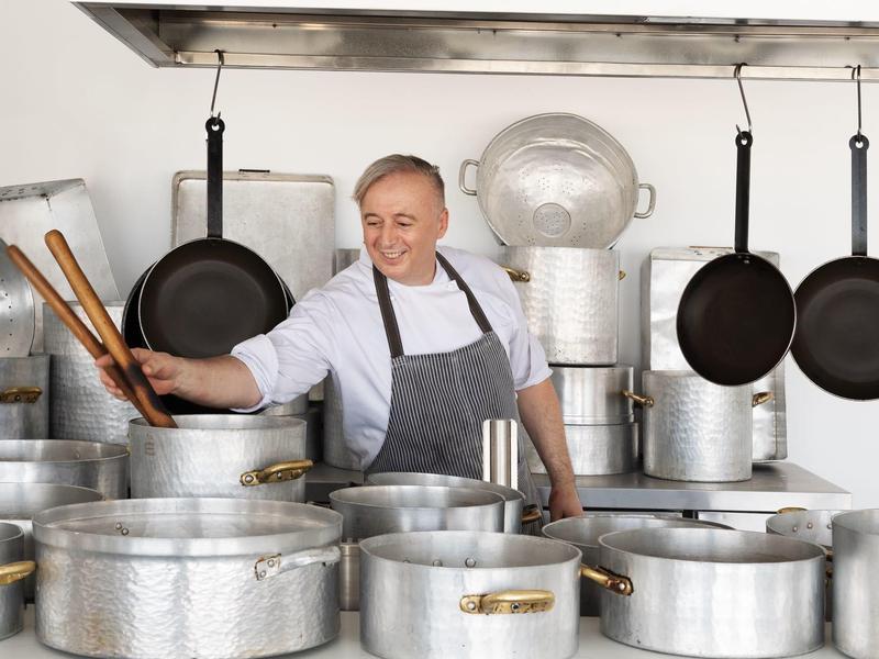 Chef standing in a kitchen with large pots and pans, holding cooking utensils.