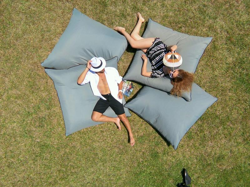 Two people relaxing on large gray floor cushions on a grassy lawn.