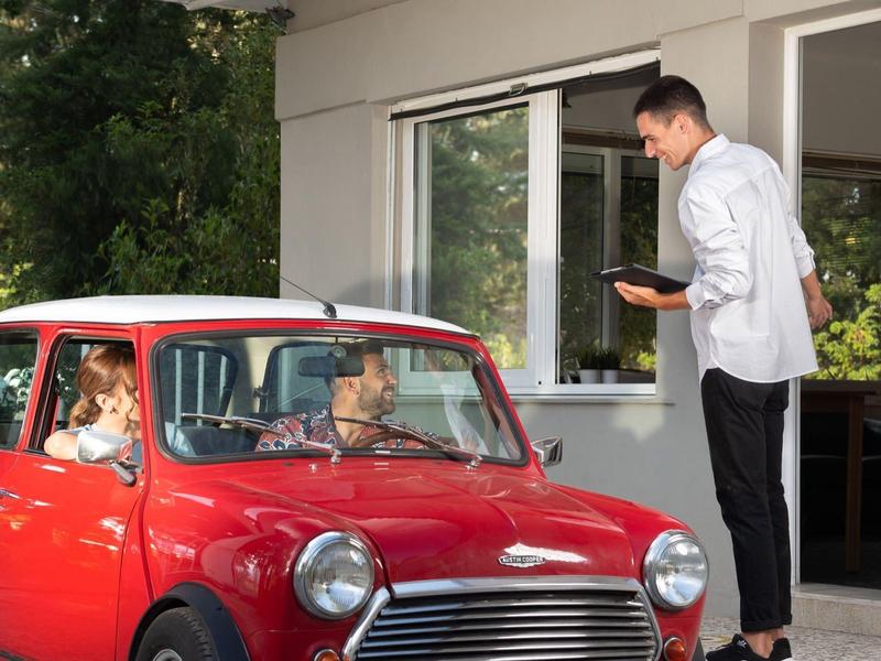 A man in a white shirt picks up food from a window to a red car with two people inside.