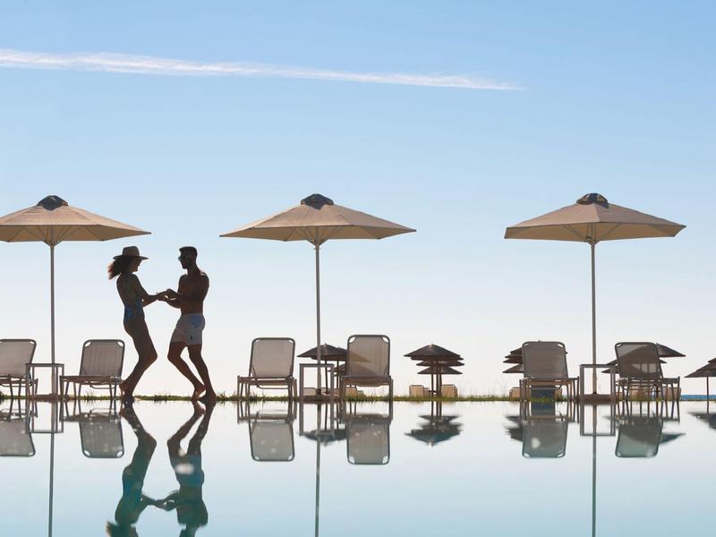 Couple dancing by pool with loungers and umbrellas, clear sky in the background.
