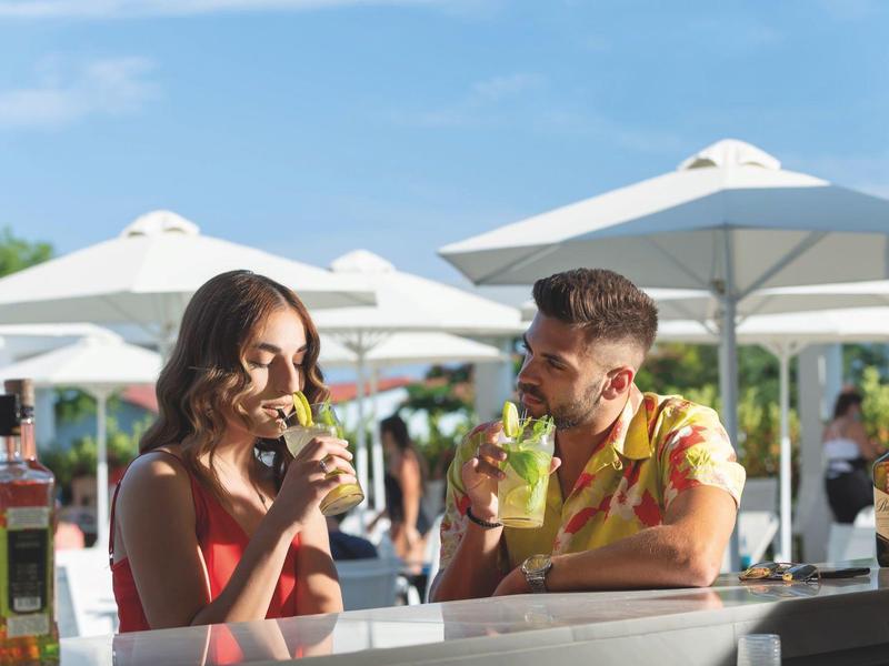 Couple enjoying cocktails at an outdoor bar under umbrellas in daylight.