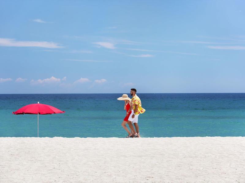 A couple walks on the beach with a red umbrella and blue sea in the background.