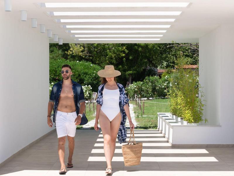 A couple in swimwear walking through a bright, modern passage with stripes of sunlight.