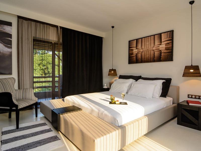 Modern hotel room with double bed, striped bench, chairs, and balcony with forest view.