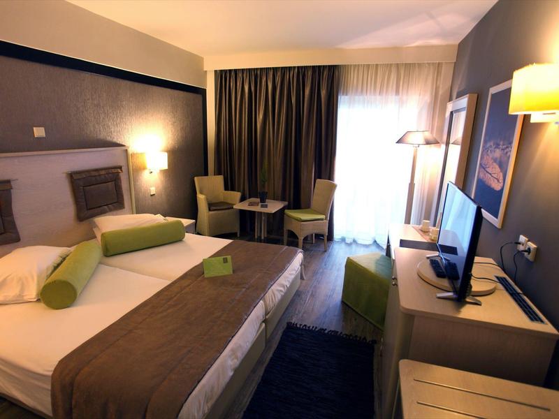 Modern hotel room with double bed, chairs, desk, TV, and a window with dark curtains.