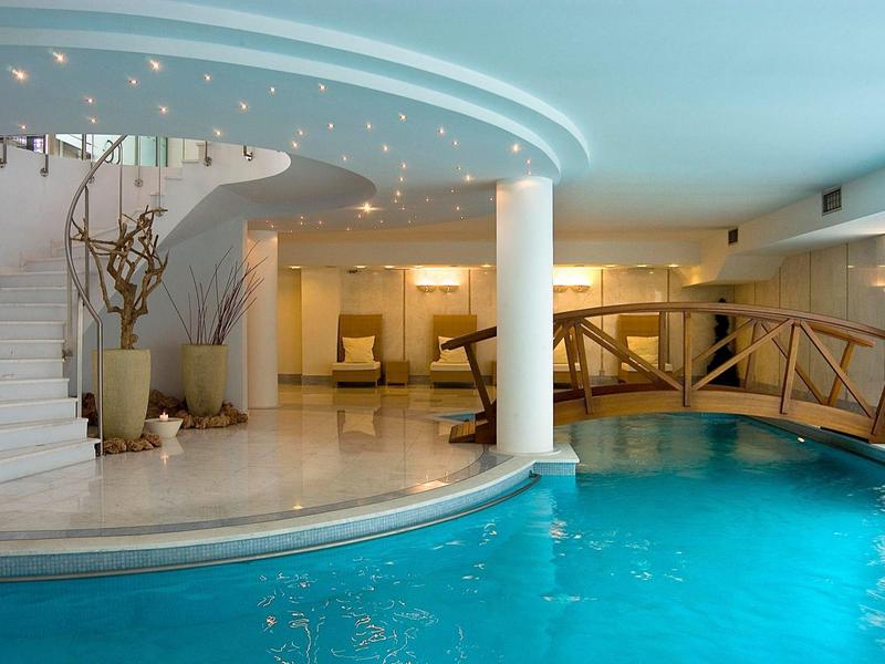 Indoor hotel pool with curved stairs, wooden bridge, and modern seating area.