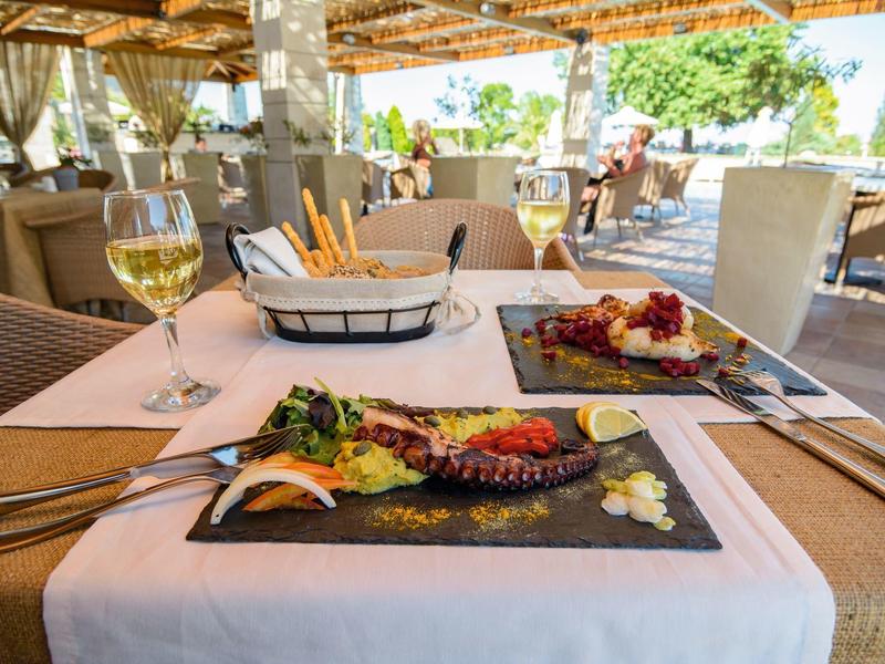 Outdoor dining setup with wine, breadsticks, and gourmet dishes on a restaurant table under shaded patio.