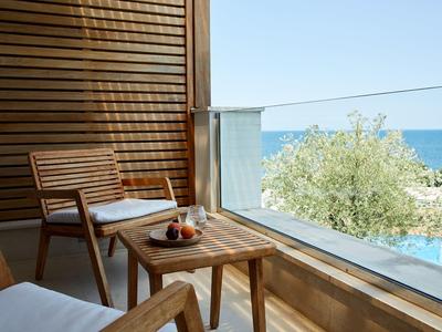 Balkon mit Holzstühlen, Tisch, Blick auf Meer und Baum, helle Farben, sonniger Tag.