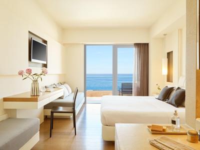 Helles Hotelzimmer mit Doppelbett, Schreibtisch, Bildschirm und Meerblick durch große Fenster.