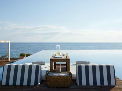 Zwei gestreifte Liegen stehen auf Holzterrasse vor einem Infinity-Pool mit Blick auf ruhiges Meer.