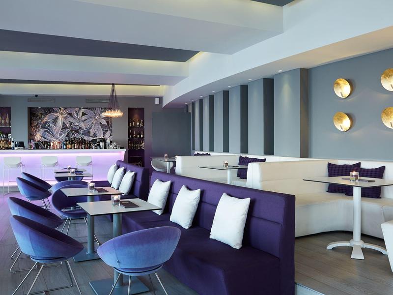 Modern eingerichtete Bar mit lila Sofas, blauen Sesseln und runden Tischen in hellem Raum.