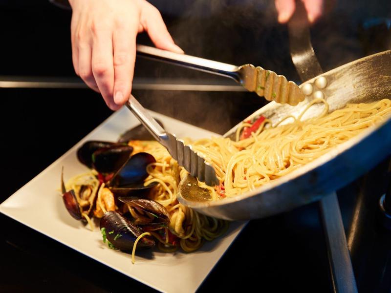 Person hebt mit Zange Spaghetti aus Pfanne auf weißen Teller mit Muscheln und Gemüse.