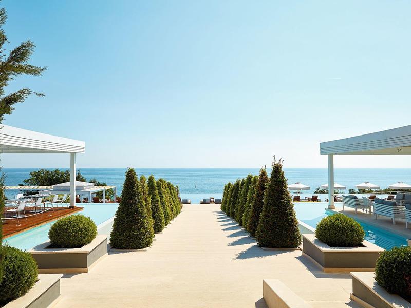 Helles Hotelzimmer mit Doppelbett, Schreibtisch, Bildschirm und Meerblick durch große Fenster.