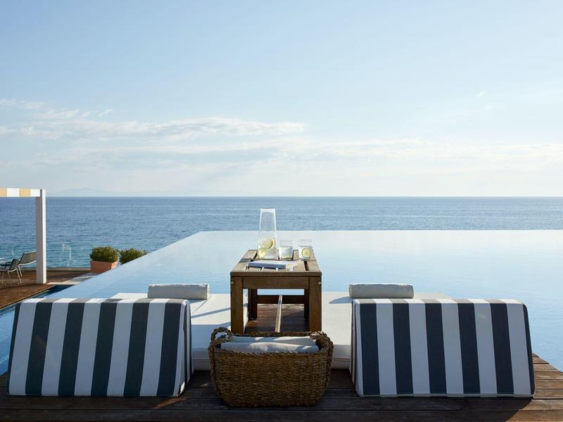 Zwei gestreifte Liegen stehen auf Holzterrasse vor einem Infinity-Pool mit Blick auf ruhiges Meer.