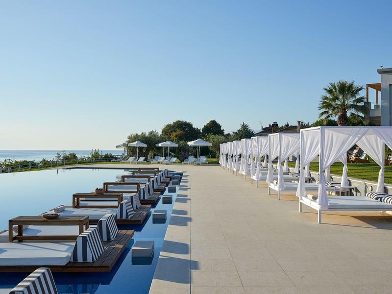 Pool mit Liegen und weißen Vorhängen neben Meer und blauem Himmel an einem sonnigen Tag.