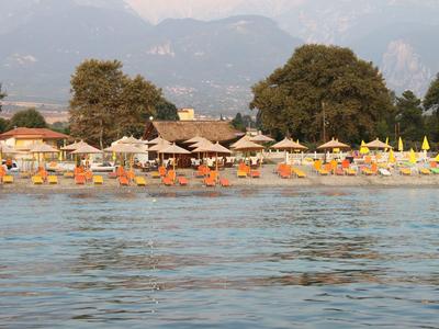 Strand mit Sonnenschirmen, Liegestühlen und Bäumen vor Bergen am Wasser.